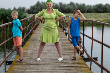 slender beautiful woman in green dress stands on bridge over river with her sons. Bright holiday clothes