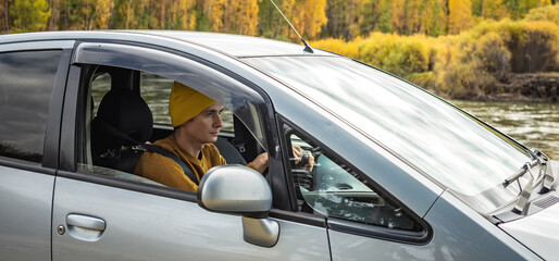 Obraz premium Man is driving a car along the river and the beautiful autumn forest. Concept of nature, travel and autumn mood