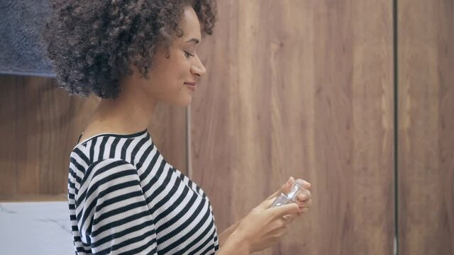 Pretty curly-haired  woman spraying perfume, smiling in bathroom mirror, glamour