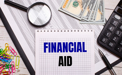 On the office table there is a notebook with the text FINANCIAL AID, a pen, a calculator, dollars, multi-colored paper clips and a magnifying glass. Stylish workplace. Business concept