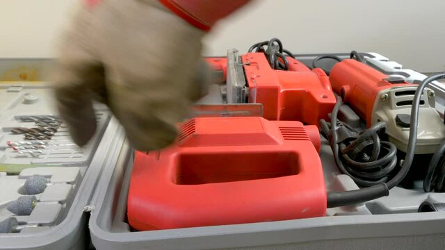 A gloved hand opening a plastic box lid then removing an electric jigsaw &ndash; with jagged blade used for cutting irregular shapes &ndash; from an assortment of power tools and attachments.