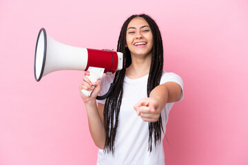 Teenager girl with braids over isolated pink background holding a megaphone and smiling while pointing to the front