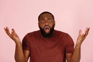 Confused young African-American person in brown t-shirt shrugs looking into camera on light pink background in studio closeup