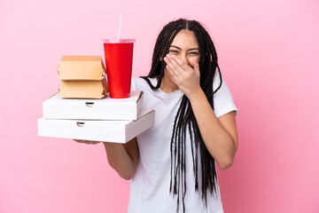 Teenager girl with braids holding pizzas and burgers over isolated pink background happy and...