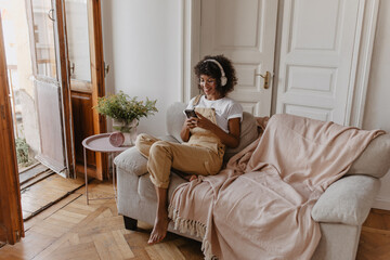 amazing young woman browses social networks on phone while sitting on couch in living room. radiant brunette with afro hair wearing headphones, glasses and casual clothes.