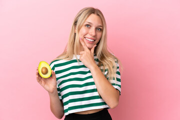 Young caucasian woman holding an avocado isolated on pink background happy and smiling