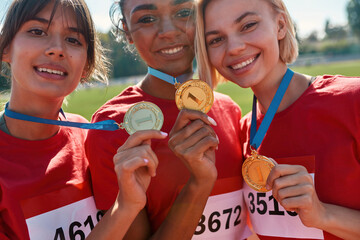 Portrait of excited diverse young female athletes showing their gold medals while standing together...