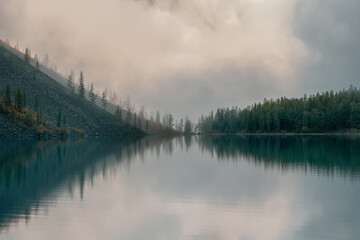 Soft focus. Silhouettes of fir hillside along mountain lake in dense fog. Reflection of coniferous trees in blue water. Alpine tranquil landscape at cool early morning. Ghostly atmospheric scenery.