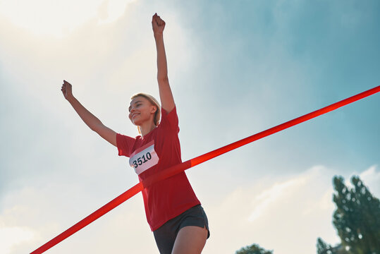 Low angle view of happy young female runner with arms raised reaching the finish line at track field during marathon outdoors