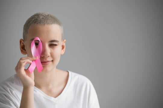 Young Woman With Short Hair Wearing A White T-shirt Holding A Pink Ribbon As A Symbol Of Breast Cancer On A White Background.