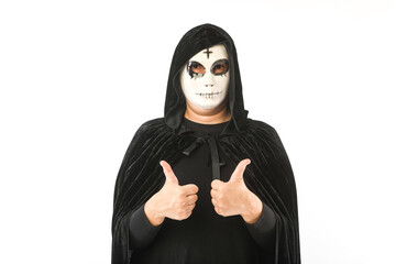 Person wearing white mask with cross on forehead and black velvet cloak with hood, raising thumbs up, on white background. Carnival, Halloween and Day of the Dead concept.