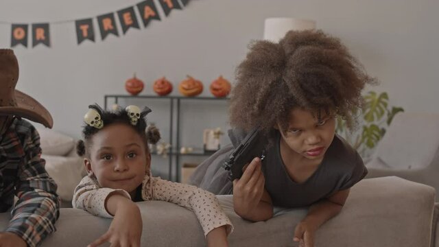 Slowmo tracking shot of playful African-American school boys and girls in Halloween costumes having fun making spooky faces on camera, posing with toy gun, vampire teeth and other decor