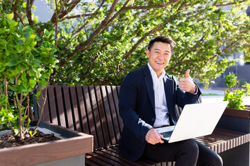 Cheerful asian man looking at camera and smiling businessman working on laptop sitting on park bench near office on break