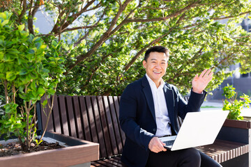 Cheerful asian man looking at camera and smiling businessman working on laptop sitting on park bench near office on break
