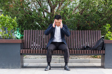 exhausted young asian business man office worker sitting on bench in city park outdoors. stressed overworked male with a headache massages head. Depressed Tired employe suffering pain sick work