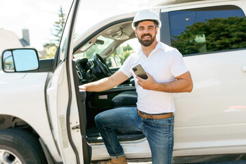 Man engineer builder wearing a white hard hat, shirt in front of his pickup using cellphone - obrazy, fototapety, plakaty