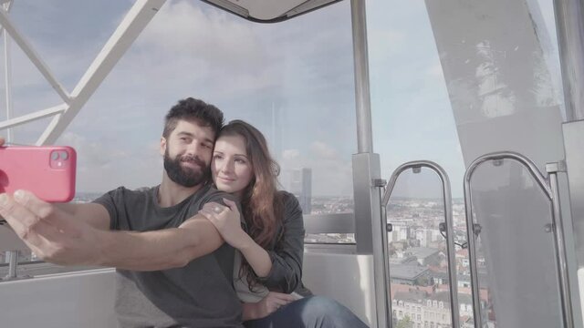 Young couple in love sitting together on ferris wheel spinning, taking selfies and skyline pictures using mobile phone, young adults traveling and visiting Brussels Europe travel destination