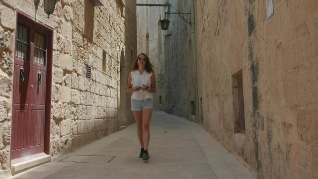 Beautiful Woman In Sunglasses Walking Alone In An Alley Through Old Buildings In Mdina, Malta. static