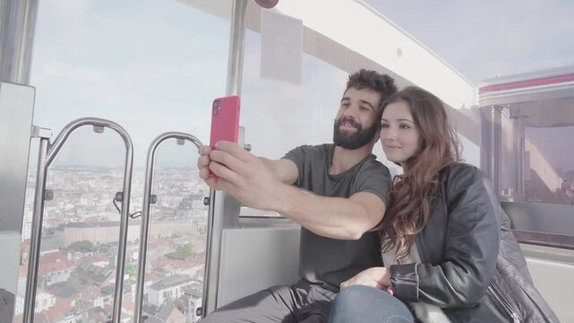 Romantic young couple traveling sitting together on ferris wheel, taking selfies and skyline pictures using mobile phone, young adults traveling and visiting Brussels Europe travel destination
