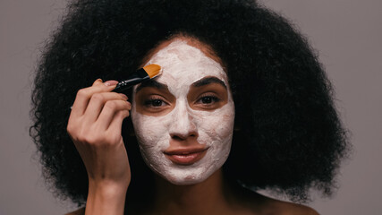 african american woman looking at camera while applying clay mask isolated on grey