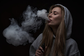 Portrait of beautiful blonde girl in gray hoodie smokes vape isolated on black studio background,...