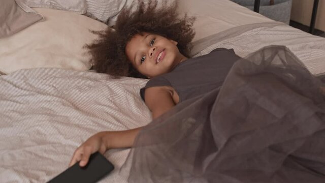 Slowmo shot of playful 8-year-old African-American girl in big dress lying down on bed having fun making funny faces on smartphone