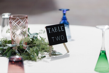 White table tablecloth blue glasses upside-down and pink vase on the wedding Table two written on black card stands near green floral decor in the center of dinner table  Table Number two