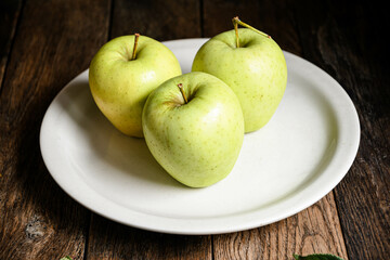 Three green apples on a white ceramic plate, on a wooden table.