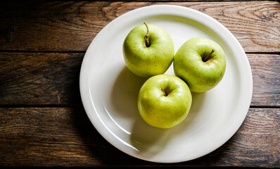 Three green apples on a white ceramic plate, on a wooden table.