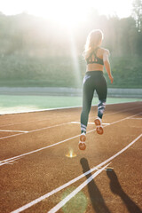 Obraz premium Rear view of athletic woman running on track stadium at summer morning light. Fit woman running on racetrack during training session. Female runner practicing on athletics race track. Sportswoman