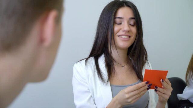 Young woman with long hair and manicure wearing white jacket sitting in class and using colorful cards, talking to man on blurred foreground. Fellow students studying together and using handouts