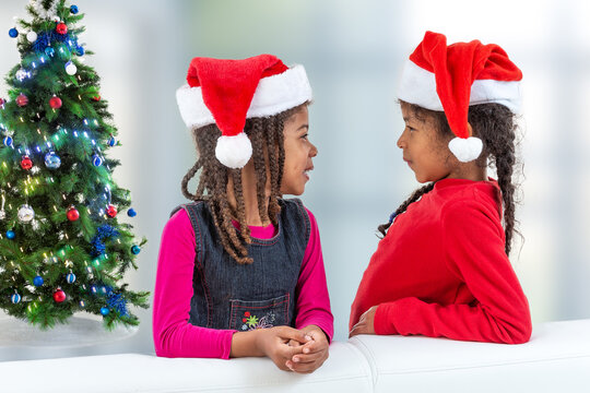 Two Happy Little Girl In Santa Hat, Stylish Sister, Happily Looking At Each Otherto Camera While Standing Near Chritsmas Tree On White Background, Profile View.