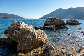 Fototapeta na wymiar Big rocks in crystal clear azure water with green hills on horizon, coast of Lefkada island in Greece. Summer nature travel to Ionian Sea