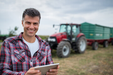 Portrait of happy farmer holding tablet standing in front of tractor with trailer.