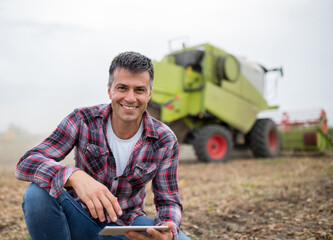 Attractive young farmer holding tablet crouching in front of harvester in soy field