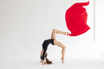slim artistic teenager girl in black leotard trains on white background with red fabric in her...