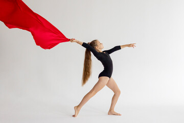 slim artistic teenager girl in black leotard trains on white background with red fabric in her hands in rhythmic gymnastic exercise, children's professional sports