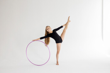 Obraz na płótnie Canvas slim artistic teenager girl in black leotard trains on white background with hoop in her hands in rhythmic gymnastic exercise, children's professional sports