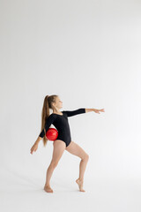 Fototapeta na wymiar slim artistic teenager girl in black leotard trains on white background with red ball in her hands in rhythmic gymnastic exercise, children's professional sports