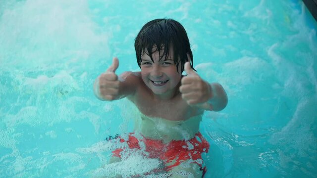 Joyful boy showing thumbs up smiling looking at camera standing in bubbling azure blue water in swimming pool. Portrait of hyperactive happy Caucasian child posing outdoors on summer vacations