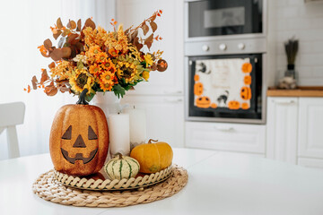 A vase of flowers,a jack pumpkin and candles on a tray. In the background - the interior of a white kitchen in Scandi style. The concept of home and comfort. Autumn decor for the Halloween holiday.