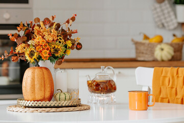 Still-life. Yellow, orange flowers in a vase, pumpkins, a teapot and a cup of tea on a white table...