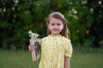 Little girl in a yellow dress stands with a book and wildflowers in nature in the field