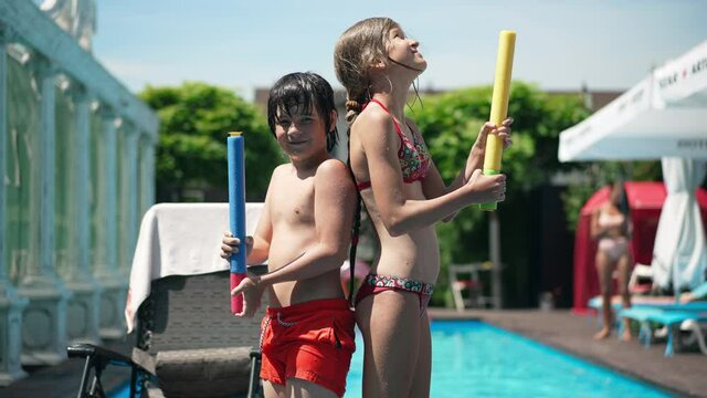 Side view of confident Caucasian children standing at swimming pool with water gun back to back and looking at camera. Cute boy and girl posing in sunshine on summer resort outdoors. Friendship