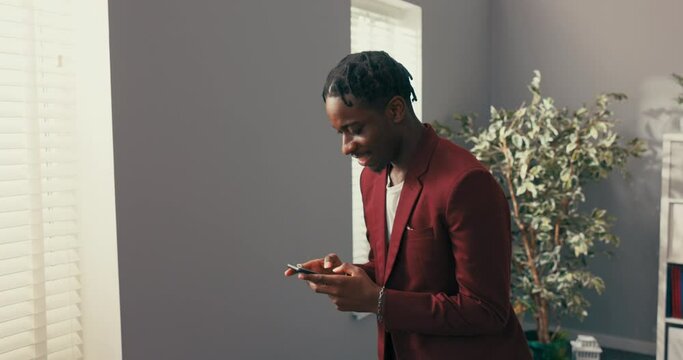 The silhouette of dark-skinned man, businessman, boss, company director, dressed in maroon suit, standing at window covered by blind, holding phone in hands, answering messages, wonders