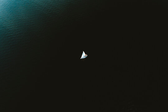 Aerial view of tiny yacht with white sails floating in calm ocean with no wind.