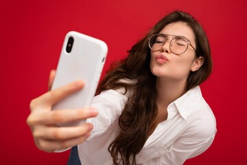 Closeup photo of sexy beautiful positive young brunette woman wearing white shirt and optical glasses isolated over red background holding in hand and using mobile phone taking selfiie looking at