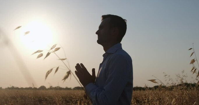 A man stands in profile facing the sun, arms crossed in front of him in boats, a man prays while standing in a wheat field.