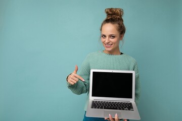 Photo of beautiful happy dark blond young woman with gathered curly hair looking at camera pointing finger at netbook holding computer laptop with empty monitor screen with mock up and copy space