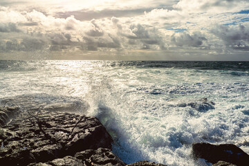 Powerful wave crushes against ragged stone coast creating splash of water. Dark dramatic light....